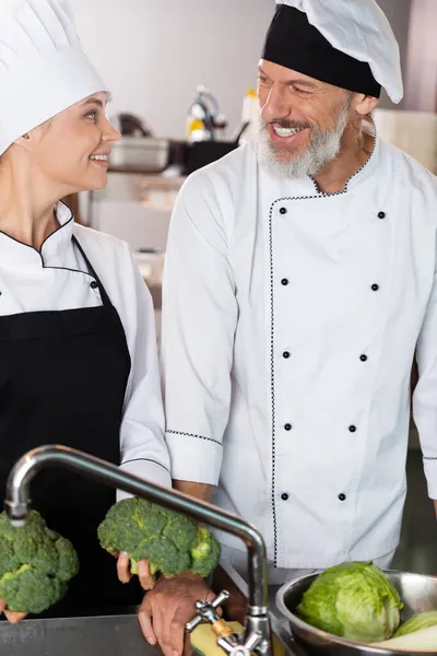 Chefs positivos sosteniendo verduras cerca del grifo en la cocina del restaurante - foto de stock
