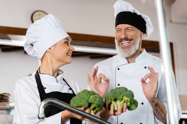 Chef sonriente mostrando buen gesto cerca de colega con brócoli en la cocina - foto de stock