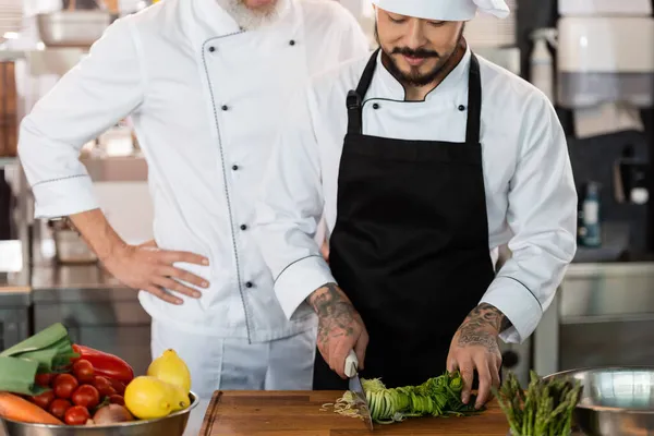 Chef asiático cortando puerro cerca de colega y verduras en cocina — Stock Photo