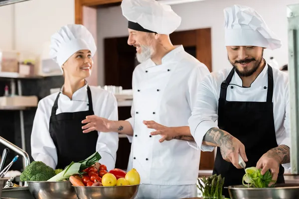 Sorrindo asiático chef segurando em fatias de alho-porro enquanto colegas falando na cozinha — Fotografia de Stock