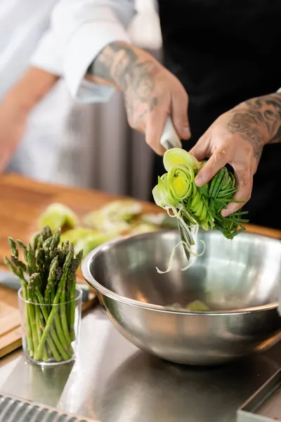 Vista cortada do chef segurando alho-porro fatiado e faca perto de tigela e espargos na cozinha — Fotografia de Stock