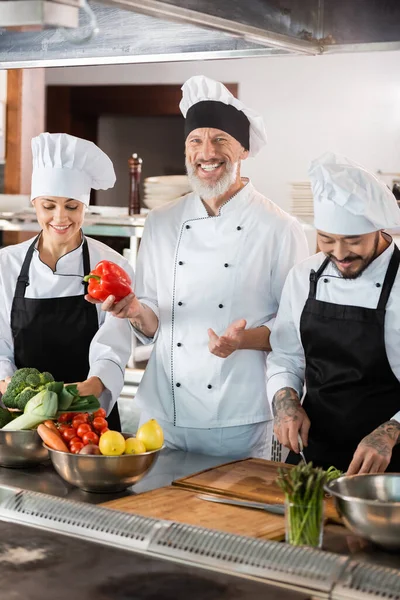 Chef feliz sosteniendo pimiento cerca de colegas interracial y verduras en la cocina - foto de stock