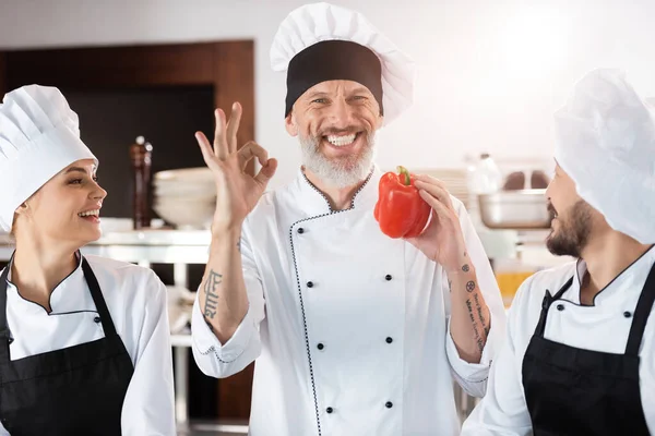 Chef sonriente mostrando ok y sosteniendo pimiento cerca de colegas multiétnicos en la cocina - foto de stock