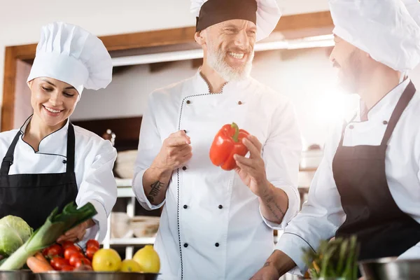 Chef apuntando a pimiento cerca sonriente asiático colega en restaurante cocina - foto de stock