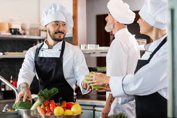 Lächelnder asiatischer Koch gibt Kollegen in Restaurantküche Gemüse — Stockfoto