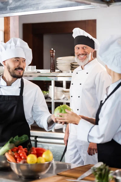 Positive chef looking at interracial colleagues with vegetables in kitchen — Stock Photo