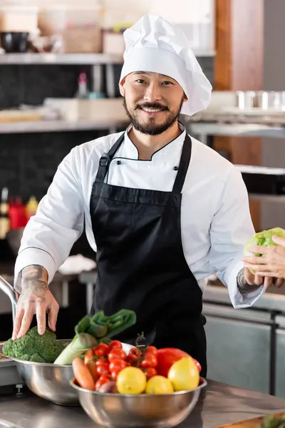 Lächelnder asiatischer Koch holt Gemüse aus Schüssel bei Kollegin in Küche — Stockfoto