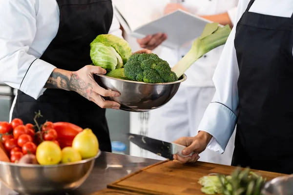 Vista ritagliata dello chef tatuato che tiene ciotola con verdure vicino al collega con coltello in cucina — Foto stock