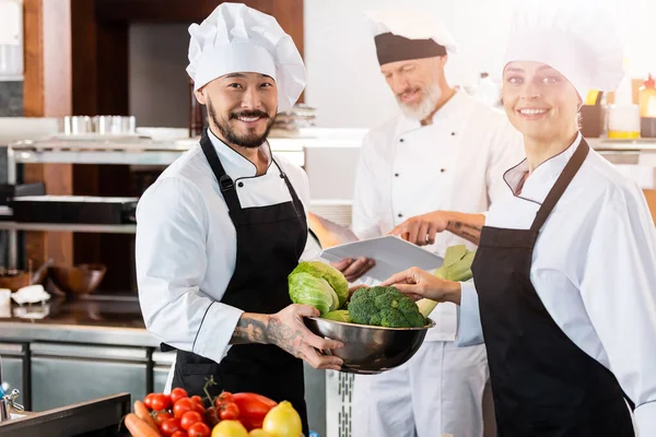 Positive gemischtgeschlechtliche Köche mit frischem Gemüse schauen in die Kamera neben Kollegen mit Kochbuch in der Küche — Stockfoto