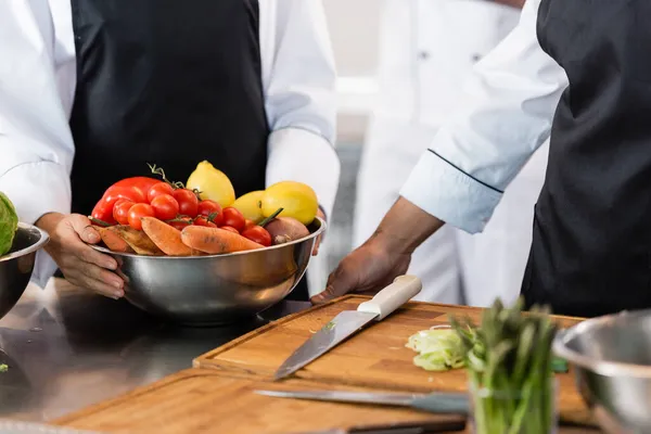 Vue recadrée du chef tenant un bol avec des légumes mûrs près d'un collègue et des planches à découper dans la cuisine — Photo de stock