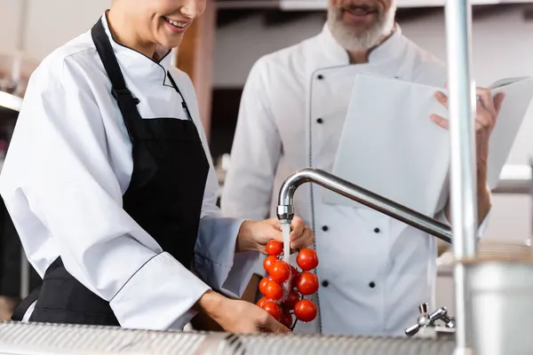 Cropped view of chef washing cherry tomatoes near colleague with cookbook in kitchen — Stock Photo