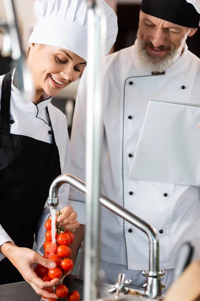 Lächelnder Koch wäscht in Restaurantküche Kirschtomaten neben Kollegin mit Kochbuch — Stockfoto