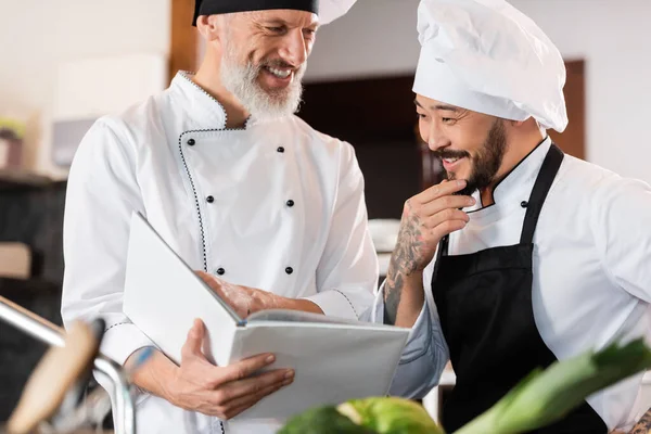 Chefs inter-raciais alegres trabalhando com livro de receitas na cozinha — Fotografia de Stock