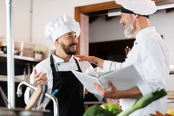 Positive chef holding cookbook while talking to asian colleague near faucet and vegetables in kitchen — Stock Photo