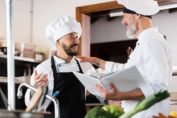 Chef positivo que prende o livro de receitas ao falar com colega asiático perto da torneira e legumes na cozinha — Fotografia de Stock