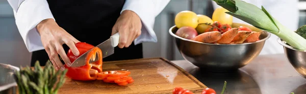 Vista recortada del chef cortando pimiento cerca de verduras maduras en la cocina, pancarta - foto de stock