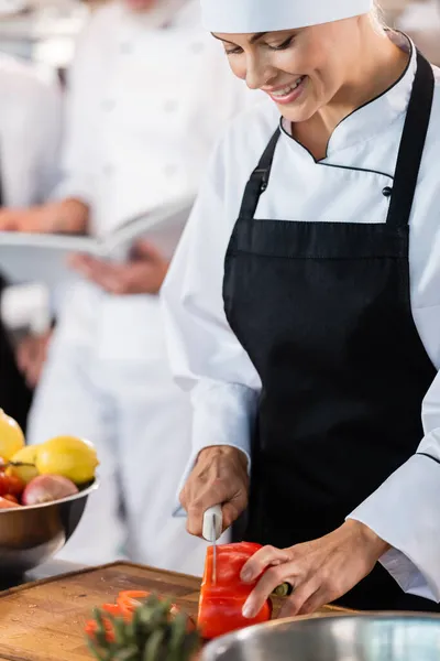 Chef positivo em avental de corte de pimentão a bordo na cozinha — Fotografia de Stock