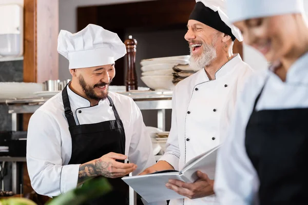 Fröhliche gemischtrassige Köche reden, während sie in der Küche Kochbuch in der Nähe verschwommener Kollegen halten — Stockfoto