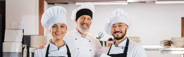 Улыбающийся шеф-повар обнимает межрасовых коллег в шапочках на кухне, баннер — стоковое фото