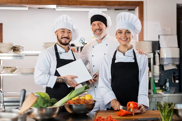 Chef asiático segurando livro de receitas perto de colegas sorridentes e legumes na cozinha — Fotografia de Stock