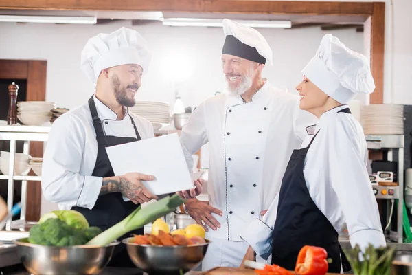 Asian chef holding cookbook and talking to smiling colleagues near food in kitchen — Stock Photo