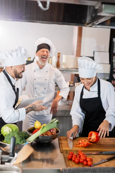 Asian chef holding cookbook near laughing colleagues and fresh food in kitchen — Stock Photo