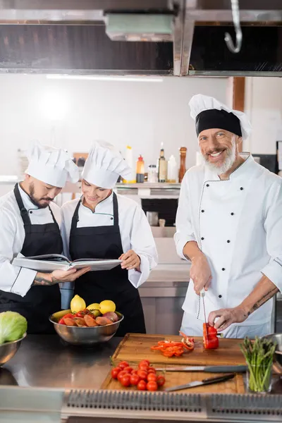 Sonriente cocinero maduro cortando pimiento cerca de colegas interracial leyendo libro de cocina en cocina — Stock Photo