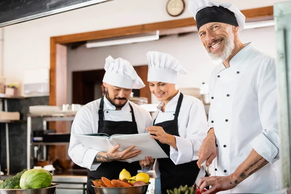 Positive chef holding knife near multiethnic colleagues in aprons reading cookbook and vegetables in kitchen — Stock Photo