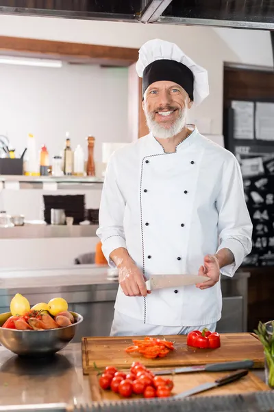 Улыбающийся шеф-повар держит нож рядом с спелыми овощами и разделочными досками на кухне — стоковое фото