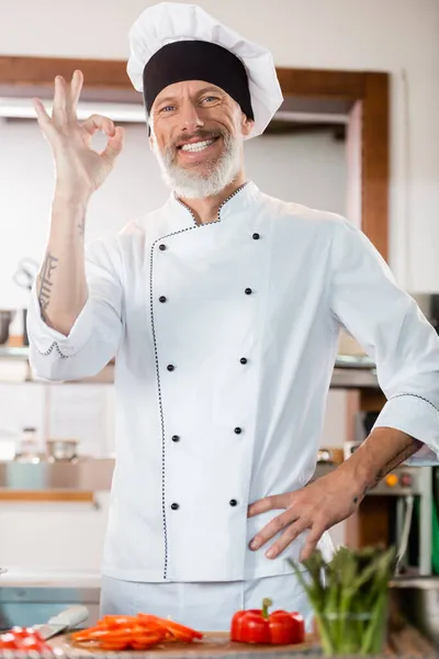 Chef positivo mostrando buen gesto cerca de verduras borrosas en la cocina - foto de stock
