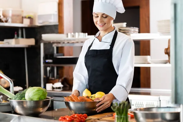 Chef sonriente en delantal mirando verduras maduras en la cocina del restaurante - foto de stock