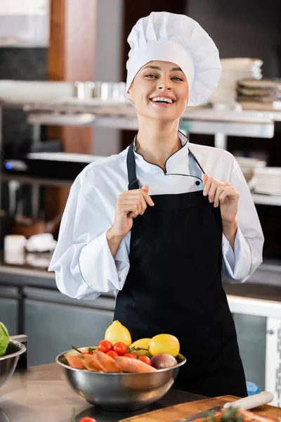 Chef positivo tocando delantal y mirando a la cámara cerca de verduras frescas en la cocina - foto de stock