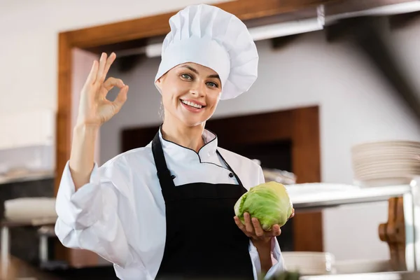 Smiling chef showing ok gesture and holding cabbage in kitchen — Stock Photo