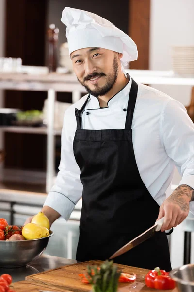 Positivo asiático chef celebración cuchillo cerca de verduras frescas y tabla de cortar en la cocina - foto de stock