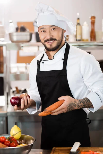 Позитивный азиатский шеф-повар держит органические овощи рядом с разделочной доской на кухне — стоковое фото