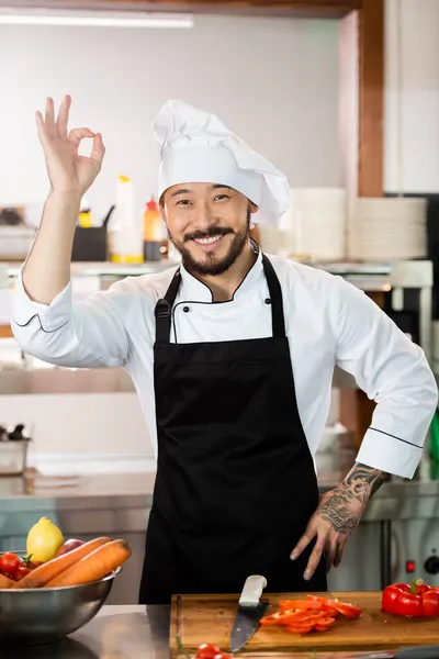 Sonriente asiático chef mostrando bien gesto cerca de la tabla de cortar y verduras en la cocina - foto de stock