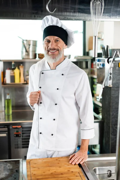 Positiver Koch in Uniform blickt in die Kamera in der Nähe von Schneidebrett und Herd in der Küche — Stockfoto