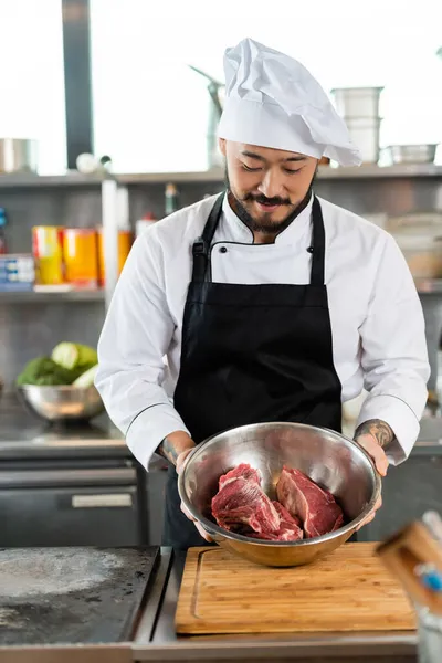 Asian chef in apron and hat holding bowl with raw meat in kitchen — Stock Photo