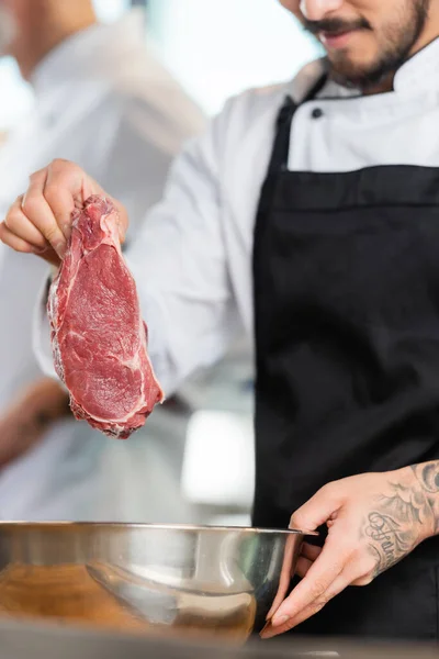 Cropped view of tattooed chef holding raw meat near bowl in kitchen — Stock Photo