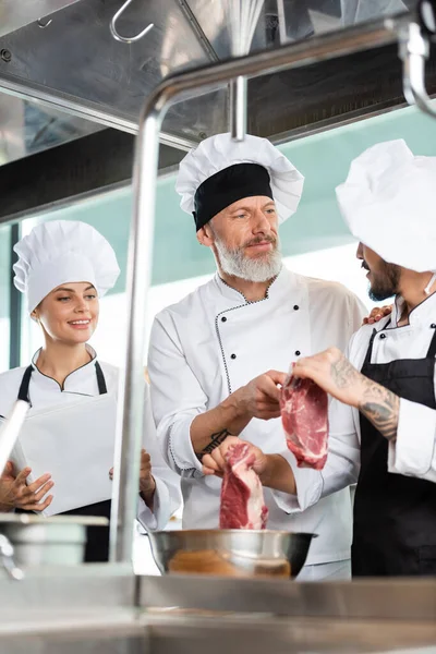 Chef señalando carne cruda cerca de colegas con libro de cocina en la cocina — Stock Photo