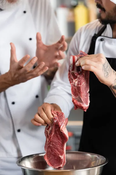 Cropped view of chef holding raw meat near bowl and colleague in kitchen — Stock Photo