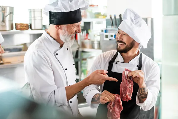 Sorridente chef indicando carne cruda vicino tatuato asiatico collega in cucina — Foto stock