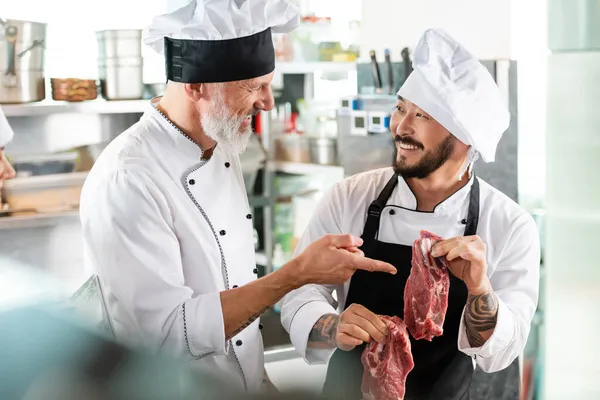 Lächelnder Koch zeigt in der Küche neben tätowiertem asiatischen Kollegen auf rohes Fleisch — Stockfoto