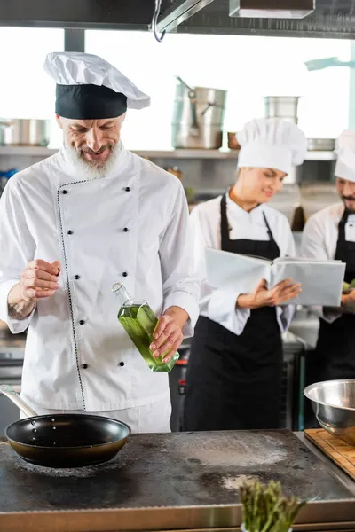 Chef sorridente segurando azeite perto da frigideira na panela perto de colegas inter-raciais borrados com livro de receitas na cozinha — Fotografia de Stock