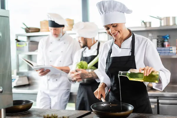 Усміхнений шеф-кухар в фартусі заливає оливкову олію на сковороді біля розмитих колег з кулінарною книгою на кухні ресторану — стокове фото