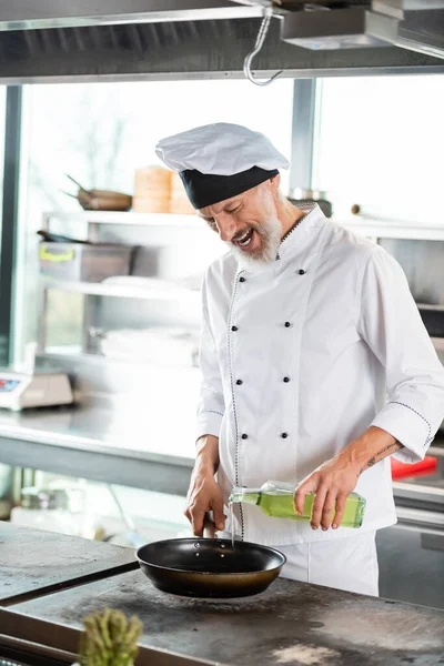 Cuoco maturo positivo che versa olio d'oliva su padella sul piano cottura in cucina — Foto stock