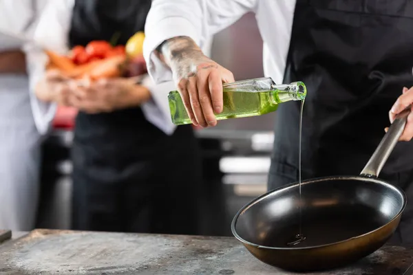 Cropped view of tattooed chef pouring olive oil on frying pan in kitchen — Stock Photo