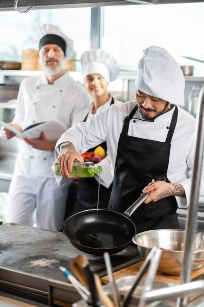 Chef asiatique verser de l'huile sur la poêle près des ustensiles de cuisine et des collègues flous avec livre de cuisine dans la cuisine — Photo de stock