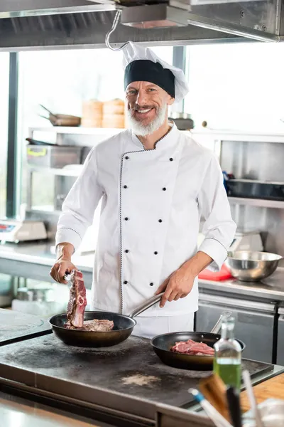 Chef sorridente em uniforme cozinhar carne no fogão na cozinha do restaurante — Fotografia de Stock