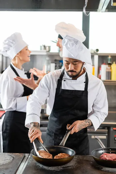 Asiatischer Koch bereitet Fleisch auf Herdplatte zu, während verschwommene Kollegen in der Küche reden — Stockfoto